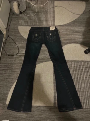 True Religion Joey Low Rise Flare jeans - Snygga mörkblå jeans från True Religion, modell Joey Low Rise Flare. De har låg midja, utställda ben och klassiska kontrastsömmar. Bakfickorna har lock och dekorativa detaljer med metallknappar. Perfekt för dig som gillar retrovibbar och coola jeans. Om du är osäker på om detta är rätt storlek för dig så är jag 166cm lång och dom passar perfekt på mig.