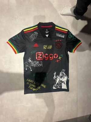 Ajax x Bob Marley t-shirt Adidas M - Svart Ajax x Bob Marley t-shirt från Adidas med färgglada detaljer i rött, gult och grönt på ärmar och axlar. Tryck med Bob Marley, citat och graffitistil, samt Ziggo-logga. Tillverkad i lätt och ventilerande AEROREADY-polyester.