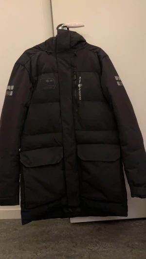 Svart dunjacka från Sail Racing - Svart dunjacka från Sail Racing med huva och stora fickor framtill. Jackan har logotyp på ärmen och bröstet, samt dragkedja och knappar. Materialet är polyester och dun, och jackan är vind- och vattenavvisande. Perfekt för kalla vinterdagar. 