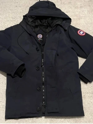 Canada Goose XS navy dunjacka  - Säljer en navy dunjacka från Canada Goose i storlek XS. Jackan har huva, dragkedja och knappar framtill, samt klassisk logga på ärmen och broderad patch på ryggen. Insidan är quiltad och fodrad med dun för extra värme. Perfekt för kalla dagar.