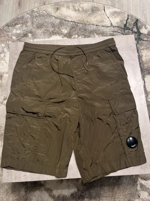 Olivgröna cargo shorts från C.P. Company - Snygga olivgröna cargo shorts från C.P. Company med ikonisk linsdetalj på benet. De har elastisk midja med snörning, flera fickor och är tillverkade i lätt polyester som ger en cool, lite glansig look. Perfekta för sommaren och streetwear-stilen. Size 50, oanvända 10/10 skick! Kvitto finns och köpta på NK i Göteborg!