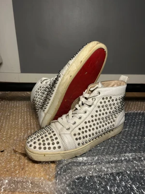 Christian Louboutin vita sneakers med nitar - Säljer ett par vita high-top sneakers från Christian Louboutin med ikonisk röd sula och silverfärgade nitar över hela skon. Skorna har snörning och är tillverkade i skinn, med rund tå och platt sula. Märkeslogga på sidan. Bra skick och skönt ingådda så väldigt bekväma. Har äkthetsbevis och kvitto som jag skickar vid intresse. Har låda, dustbags och massa andra små grejer som kom med som fås med vid köp.
