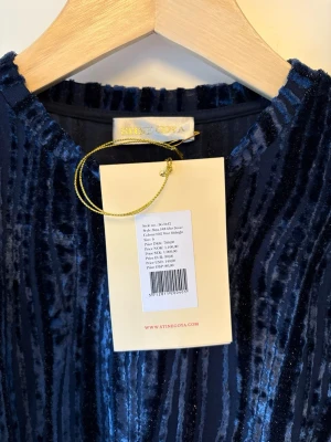 Mörkblå randig sammetsblus Stine Goya - Säljer en mörkblå blus från Stine Goya i sammetsmaterial med glittriga ränder. Blusen har lång ärm, rund hals och en slim passform. Materialet är en mix av rayon, polyester, metallisk tråd och elastan som ger en lyxig känsla och snygg struktur. Ny med lappar kvar, ej använd.