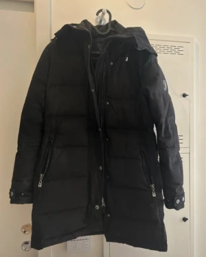 Svart lång dunjacka Hollies 36 - Svart lång dunjacka från Hollies med huva, stora dragkedjor och knappar framtill. Jackan har quiltad design, två fickor med dragkedja och snygga detaljer vid ärmslut. Perfekt för kalla dagar och har en stilren look med diskret logga på ärmen.