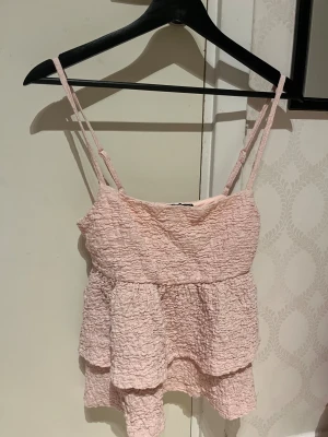 Ljusrosa volangtop från Gina Tricot - Supersöt ljusrosa topp från Gina Tricot i storlek S. Toppen har smala axelband, volangdetaljer och ett krinklat tyg som ger en cool struktur. Perfekt för dig som gillar romantisk och feminin stil. Passar till jeans eller kjol för en fräsch look.