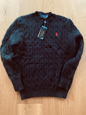 Svart kabelstickad tröja Polo Ralph Lauren - Kabelstickad tröja från Polo Ralph Lauren i storlek M. Klassisk rund hals och långärmad modell med röd broderad logga på bröstet. Tröjan är gjord i mjuk ull och har ribbade muddar vid ärmslut och nederkant. Perfekt för dig som gillar stilren och tidlös design.