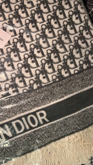 Svartvit Dior väst med logomönster - Säljer en svartvit väst från Dior med ikoniskt logomönster över hela plagget. Västen har breda ränder med DIOR-tryck och fransar längst ner för en extra touch. Materialet är stickat och känns både lyxigt och mjukt. Perfekt för dig som vill sticka ut med designerstil.