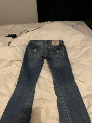 True Religion blå bootcut jeans - Säljer ett par klassiska blå jeans från True Religion med tydliga kontrastsömmar och ikoniska fickdetaljer bak. Modellen har bootcut-ben och låg midja. Jeansen är i kraftigt denimtyg och har snygga ficklock med knappar och broderade detaljer.