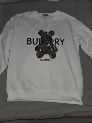 Vit sweatshirt från Burberry med nalletryck - Vit sweatshirt från Burberry med ett stort tryck av en nalle på bröstet och tydlig Burberry-logga. Tröjan har rund hals, ribbade muddar och är långärmad. Materialet är mjuk bomull, perfekt för dig som gillar streetstyle och vill sticka ut. Storlek M men passar S
