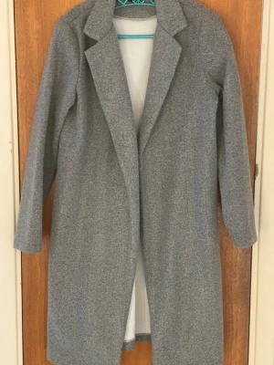 Grå lång kappa - Snygg grå kappa med klassisk krage och lång passform. Jackan har raka linjer och är helfärgad utan synliga detaljer som knappar eller bälte. Perfekt för dig som gillar stilrena och enkla plagg. Materialet känns mjukt och har en ull-liknande look.