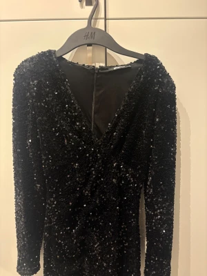 Svart paljettklänning Gina Tricot - Säljer en svart kort paljettklänning från Gina Tricot med lång ärm och v-ringning, perfekt till nyår🖤 Klänningen har en figurnära passform och dragkedja bak. Endast använd en gång!