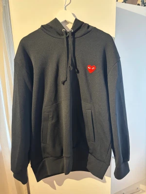 Svart CDG Play Hoodie  - Säljer nu denna snygga svarta hoodie från CDG Play då jag inte använt så mycket. Den är väldigt bra skick och storlek XL. Passar dock allt från M-XL. Nypris ligger runt 2500kr. Hör av dig om du har några frågor eller vill se fler bilder.