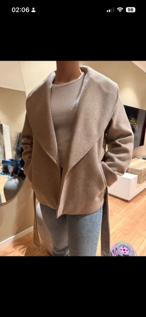 Beige omlott kappa från Vero Moda - Snygg beige kappa från Vero Moda med bred krage och omlott-design. Jackan har ett bälte i midjan för att kunna knytas och ge en schysst siluett. Materialet är mjukt och känns lyxigt, perfekt för dig som gillar stilrena och enkla plagg.