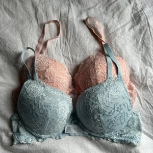 Blommiga spetsbehåar i pastell - Två söta behåar med blommig spets, en i ljusblå och en i ljusrosa. Båda har metallcharm framtill. Använt en gång. Ena är 80B och den andra 75b 