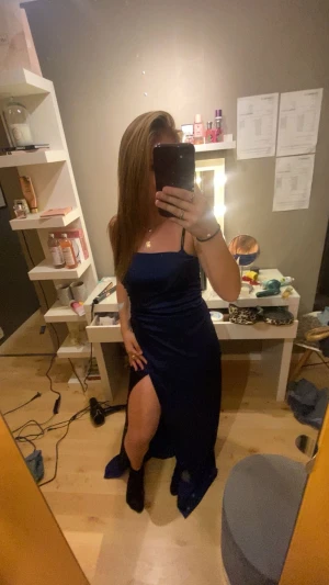 Marinblå långklänning med slits - Säljer en elegant marinblå långklänning med smala axelband och snygg slits på sidan. Klänningen har en figurnära passform och är tillverkad i ett glansigt material som ger en lyxig känsla. Perfekt för dig som vill sticka ut med en stilren look.