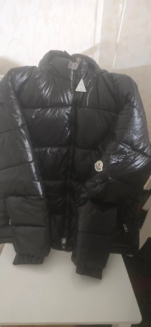 Svart Moncler pufferjacka med huva - Snygg svart pufferjacka från Moncler med glansig finish och huva. Jackan har dragkedja framtill, praktisk ficka med knapp på ärmen och ikonisk Moncler-logga. Perfekt för kalla dagar och har en modern, oversized look.