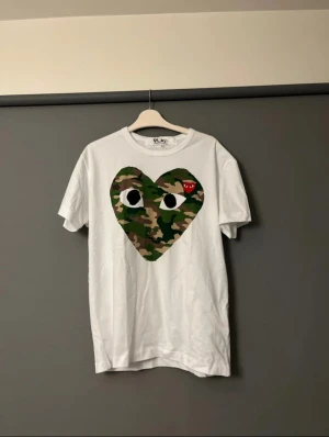 Vit Comme des Garçons Play t-shirt - Säljer en vit t-shirt från Comme des Garçons. Klassisk rund hals och korta ärmar. Perfekt för dig som har en stilren stil 
