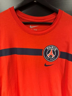 Röd PSG t-shirt från Nike XXL - Snygg röd t-shirt från Nike med Paris Saint-Germain-logga på bröstet och en marinblå horisontell rand över framsidan. perfekt för dig som gillar fotboll och vill visa ditt PSG-support.