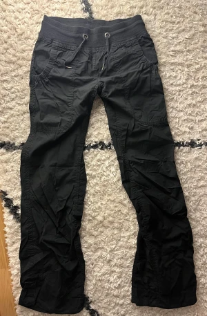 Svarta cargopats-lika byxor med snörning - Svarta cargopants-liknande byxor bred resårmidja och snörning framtill. Byxorna har sidfickor och en bakficka med dragkedja. Lösa vid benen men mer åtsittande runt midja och höft. Midwaist och långa i benen på mig som är 170