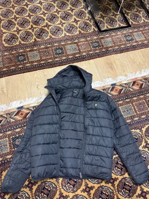Lyle scott jacka  - Svart pufferjacka från Lyle & Scott med huva och gul logotyp på bröstet. Jackan har horisontella quiltade paneler och dragkedja framtill. Perfekt för kyliga dagar och har en clean, modern look som funkar till det mesta.