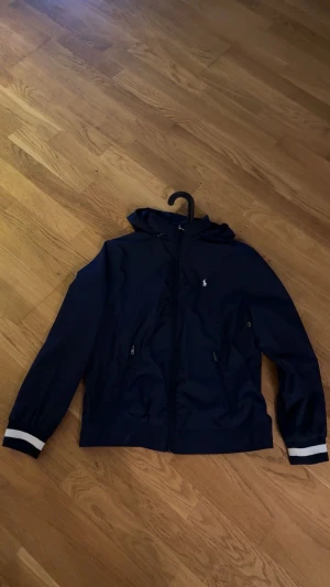 Marinblå vindjacka från Polo Ralph Lauren - Passar dig mellan 160-175 pris kan diskuteras Snygg marinblå vindjacka från Polo Ralph Lauren med huva och dragkedja framtill. Jackan har vita ränder på mudden, två fickor med dragkedja och klassisk Polo-logga på bröstet. Perfekt för dig som gillar stilren och sportig look.