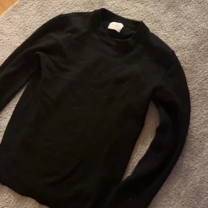 Svart merinoull tröja från a days March - En svart stickad tröja från a days march med enkel och clean design. Tröjan har lång ärm och en något högre rund halsringning. Perfekt för lager-på-lager och funkar till allt. Materialet är mjukt och skönt mot huden.