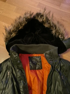 Grön bomberjacka med fuskpäls från Brave Soul - Snygg mörkgrön bomberjacka från Brave Soul med orange foder, avtagbar huva med svart foder och fuskpäls. Jackan har dragkedjor på fickorna, ribbade muddar och klassisk bomberstil. Perfekt för kalla dagar när du vill ha både stil och värme.