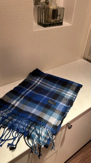 Blårutig ullväst från Burberrys - Snygg blårutig väst från Burberrys i 100% lammull, tillverkad i England. Västen har klassiskt rutmönster i blått, svart och vitt med fransar längs nederkanten. Perfekt för dig som vill ha en cool och tidlös look med premiumkänsla.