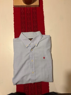 Ljusblå skjorta från Morris - Snygg ljusblå skjorta från Morris med klassisk button-down krage och broderad röd logga på bröstet. Skjortan har långa ärmar, vita knappar och är tillverkad i mjuk bomull. Perfekt för dig som gillar stilrena och tidlösa plagg med diskreta detaljer.