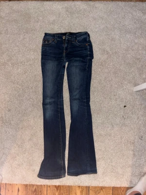 Mörkblå bootcut jeans från True Religion - Snygga mörkblå jeans från True Religion med bootcut passform och klassiska detaljer. Jeansen har dekorativa sömmar och knappar på bakfickorna. De är något slitna längst ner. Och de används inte nu pg att de är för små. 