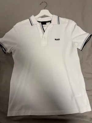 Vit polotopp från BOSS med kontrastdetaljer - Säljer en stilren vit polotopp från BOSS med svart broderad logga på bröstet. Toppen har korta ärmar och krage med svarta och grå kontrastlinjer. Tillverkad i bomull med knappslå framtill. Perfekt för dig som gillar klassisk och clean stil.