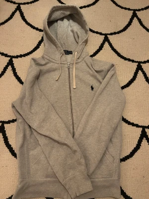 Ralph Lauren zip hoodie snören - En grå ralph lauren med zip. Den har ett blått stickat Ralph lauren märke på sin vänstra sida. den har två vita snören.