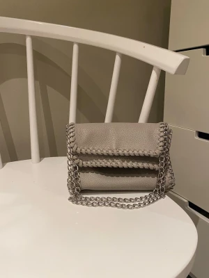 Beige axelväska med kedjedetaljer - Snygg beige axelväska i skinnimitation med  silverfärgade kedjor, axelrem och längs kanterna. Väskan har en stilren, fyrkantig form och lockstängning. Perfekt för dig som gillar en trendig och clean look. En väska som kan bäras på olika sätt.