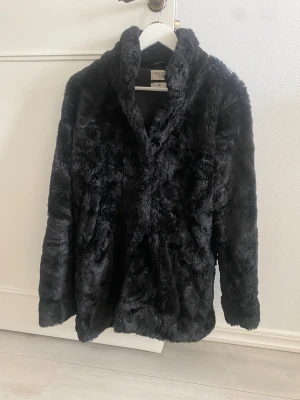 Svart pälsjacka från Tiger of Sweden - Säljer en svart pälsjacka från Tiger of Sweden i modellen Minimal. Jackan har en fluffig och mjuk yta, klassisk krage och lång ärm. Perfekt för dig som vill ha en stilren och varm look med lite lyxig känsla. Jackan är fodrad och har en rak passform.