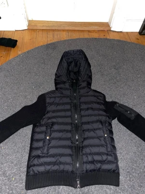 Svart Moncler dunjacka med huva - Svart dunjacka från Moncler med stickade ärmar och quiltad kropp. Jackan har huva, dragkedja framtill, fickor med tryckknappar och en extra ficka med dragkedja på ärmen. Snyggt Moncler-märke på armen och ribbade muddar vid ärmslut och nederkant.