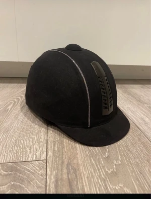 Svart ridhjälm från Kylin, storlek 52 - Snygg svart ridhjälm från Kylin med matt finish och ventiler i fram och på sidorna. Hjälmen har en klassisk rundad form, diskreta silverdetaljer längs sömmarna och justerbart hakband. Perfekt för dig som gillar ridning och vill ha en stilren look.