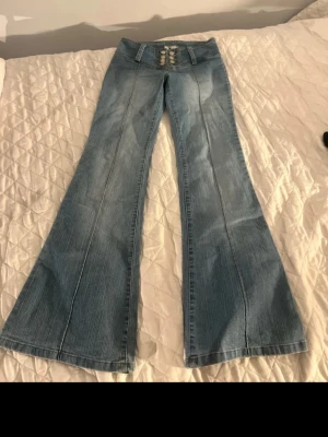 Ljusblå bootcut jeans med knappar - Snygga ljusblå bootcut jeans från K med låg midja och sex silverfärgade knappar framtill. Jeansen har markerade sömmar längs benen och är tillverkade i klassiskt denim-material. Perfekt för dig som gillar retrovibbar och vill sticka ut med coola detaljer.