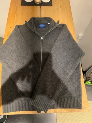 Mörkgrå stickad kofta från Jack & Jones - Mörkgrå stickad kofta från Jack & Jones med ribbad krage och muddar. Koftan har hel dragkedja framtill och är långärmad, perfekt för kyliga dagar. Materialet är mjukt och värmande, och passformen är normal. Snygg att slänga på över en t-shirt.