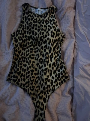 Leopardmönstrad bodysuit från H&M - Säljer en leopardmönstrad bodysuit från H&M i beige och svart. Modellen är ärmlös med hög hals och smal passform. Materialet är mjukt och stretchigt polyester som sitter snyggt mot kroppen. Den är i storlek xs.
