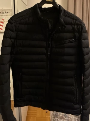 Svart pufferjacka från Belstaff - Säljer en svart pufferjacka från Belstaff med diskret logga på ärmen. Dragkedjan för bröstfickan är skadad, därav det låga priset.