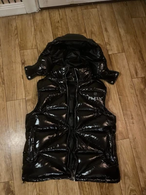 Svart pufferväst från Philipp Plein - Säljer en svart glansig pufferväst från Philipp Plein med huva och dragkedja. Västen har coola detaljer som ett stort tryck med anka och pengar på insidan samt metallbricka med logga. Perfekt för dig som vill sticka ut med streetstyle.