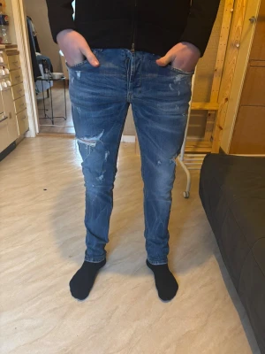 Blå dondup george slim jeans med detaljer - Säljer ett par blå slim jeans från dondup george  med slitningar och trasiga detaljer på både fram- och baksida. Jeansen har klassisk femficksdesign, låg midja och orange logga bak. Perfekta för dig som gillar en avslappnad och trendig look.
