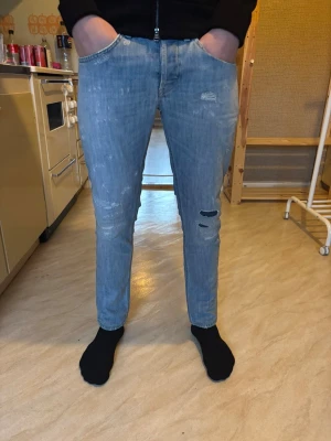 Ljusblå dondup George jeans slim fit - Säljer ett par ljusblå dondup george jeans med skinny fit. Jeansen har slitna detaljer och hål på ena benet för en trendig look. Klassisk femficksmodell med dondup logga på bakfickan. Perfekta för dig som gillar en smalare passform.