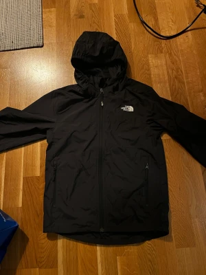 Svart vindjacka The North Face  - Svart vindjacka från The North Face med huva och dragkedja framtill. Jackan har två fickor med dragkedja och logga på bröstet samt längs ena ärmen. Tillverkad i polyester, perfekt för blåsiga dagar. OBS! Barnstorlek på jackan så egentligen Y-M i storlek