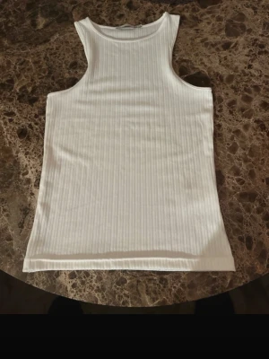 Vit ribbad linnetopp - Säljer en fräsch vit linnetopp med ribbad struktur. Toppen är ärmlös och har en klassisk rund halsringning. Perfekt för varma dagar och enkel att matcha med jeans eller shorts. Materialet känns mjukt och följsamt mot huden.