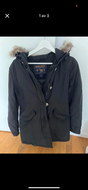 Svart parkajacka från Woolrich S - Svart parkajacka från Woolrich med huva och snygg pälsdetalj. Jackan har två stora fickor framtill, dragkedja och knappar. Materialet är polyester och dun, perfekt för kalla dagar. Klassisk och stilren modell som håller dig varm hela vintern.