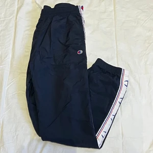 Svarta täckbyxor från Champion - Snygga svarta täckbyxor från Champion med elastisk midja och vita sidopaneler med blå och röda detaljer. Byxorna har en sportig look med diskret Champion-logga på benet och är perfekta för chill eller träning.