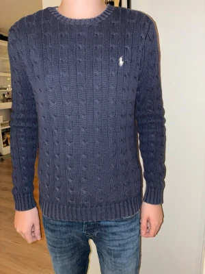 tröja Ralph Lauren - Snygg mörkblå kabelstickad tröja från Ralph Lauren med klassisk rund hals och vit broderad logga på bröstet. Tröjan är tillverkad i 100% bomull och har ribbade muddar vid ärmslut och nederkant. Perfekt för dig som gillar stilrena och tidlösa plagg.