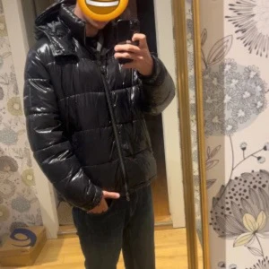 Svart pufferjacka Michael Kors L - Svart pufferjacka från Michael Kors i storlek L. Jackan har glansig yta, huva och dragkedja framtill. Quiltad design med breda paneler och diskret logga på ärmen. Perfekt för kalla dagar och har en modern, clean look.