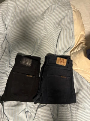Svarta och mörkblå Nudie Jeans - Två par Nudie Jeans säljes, ett par svarta och ett par mörkblå. Båda har klassisk Nudie-logga på bakfickan och läderpatch i midjan. Jeansen är raka i modellen och tillverkade i slitstarkt bomullsmaterial. Perfekta för dig som gillar stilrena och tidlösa jeans.